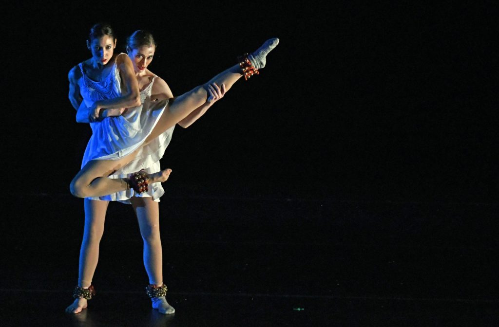 A photo still from a performance by Blue13. It shows two dancers onstage, dressed all in white. One is holding the other off of the ground, and the one being held has their leg extended up in the air.