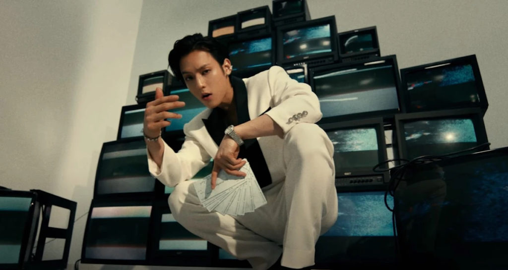 A photo still from the music video "Boom" by Huta. It shows Huta crouched in front of a bank of TV screens, looking down straight into the camera.