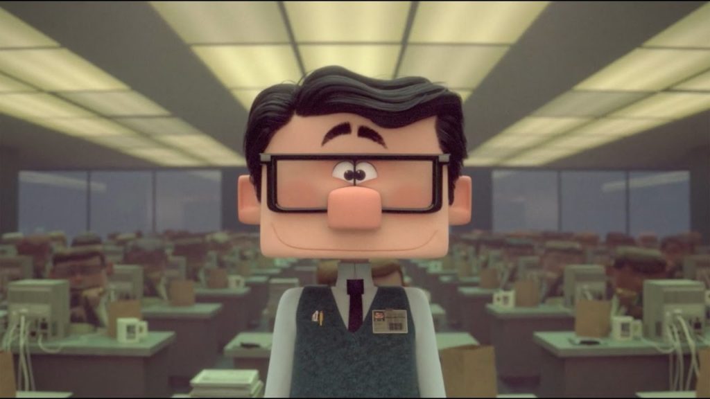 A photo still from Inner Workings. The image is of the main character, Paul, in an office, surrounded by rows of coworkers sitting in front of computers at their desks. Paul is looking straight into the camera.