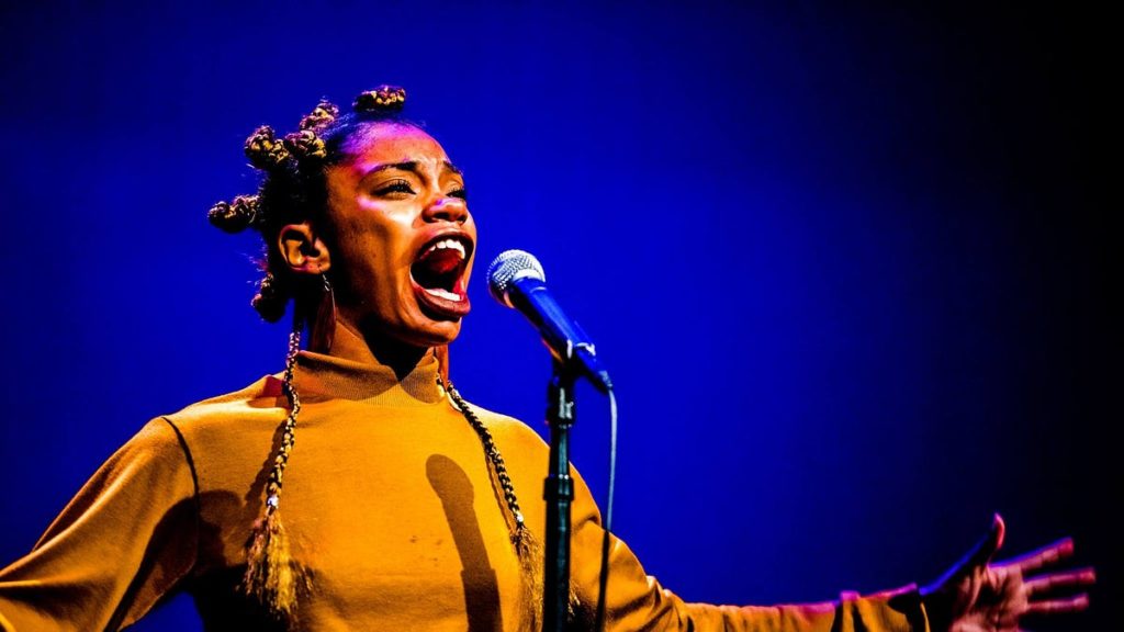 A photo of Jae Nichelle. She is standing onstage, performing into a microphone.