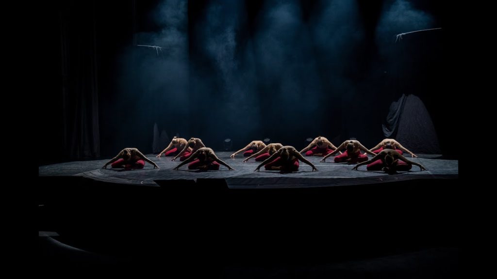 A photo of a dozen dancers onstage performing a piece by MOIRA. They are all on the ground with their heads down and arms outstretched. They are top lit with the rest of the stage in darkness.