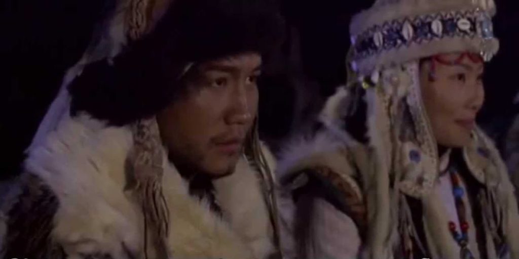 A photo still from the music video Sodura by Sarantuya. It shows a man and a woman sitting side by side in traditional Mongolian dress, looking off to the right of the camera.