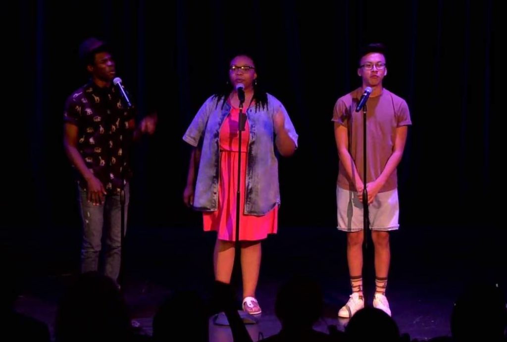 A photo of three performers, onstage, in front of microphones on stands, performing Code Switching 101.