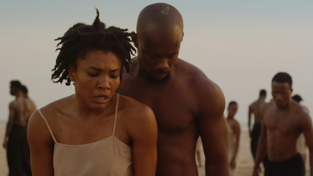 A photo still from Dancing Pina. It shows two dancers on a beach in the forefront, with the rest of the company in the background.