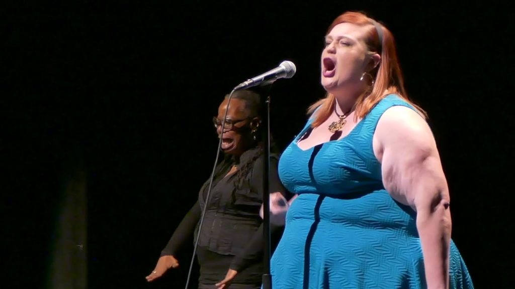 A photo of Glori B performing onstage. She is speaking directly into a microphone, and there is an ASL translator standing behind her.
