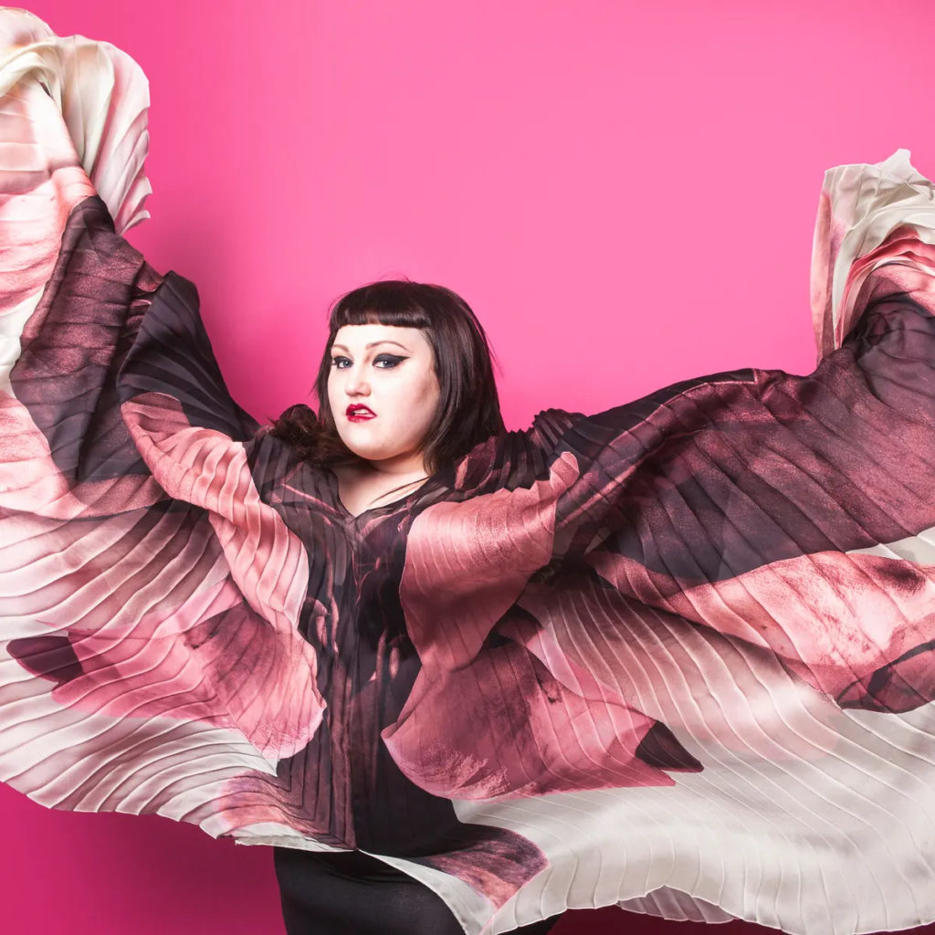 A photo of Beth Ditto, lead singer of Gossip. She's standing in front of a bright pink wall, and holding up the sides of her outfit like wings.
