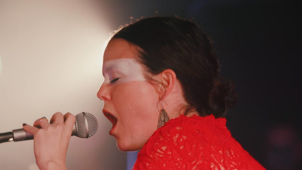 A close up photo of iskwē mid-performance. She is onstage, eyes closed, singing into a microphone.