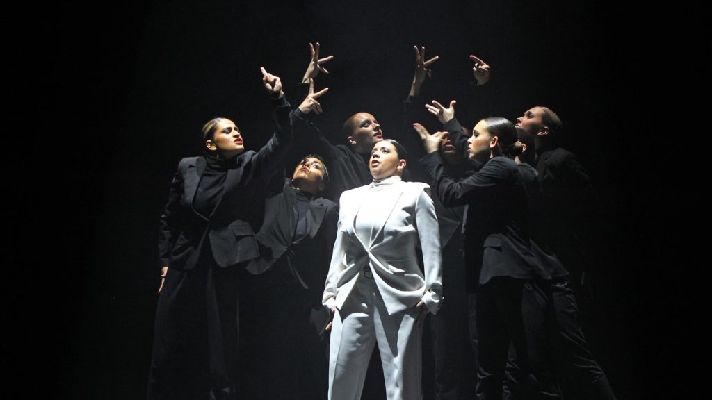 A photo still from Tendre La Main by Jennifer Romen. A dancer in a white suit is surrounded by half a dozen other dancers all in black suits and holding out their hands to the woman in white.