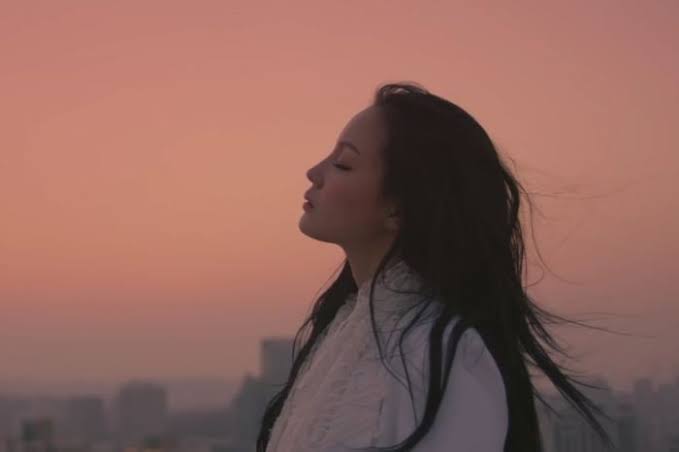 A photo of Lee Hi. She is standing in profile, eyes closed, looking up, against a sunset background.