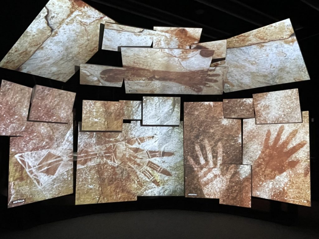 A photo of a bank of screens, artfully arranged to create a curved surface, with images of prehistoric art of hands on them.