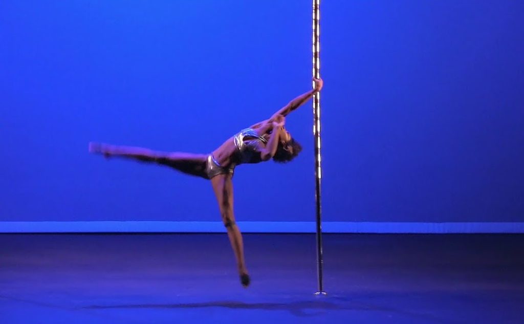 A photo of Ashley Fox mid-performance on a pole in front of a blue backdrop. She is holding on to the pole with one hand, with her legs and body extended out to the side.