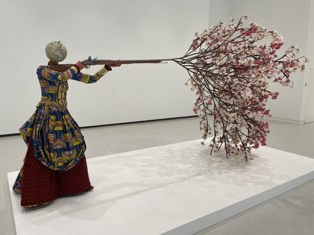 A photo of Woman Shooting Cherry Blossoms by Yinka Shonibare. It depicts a woman figure with a globe for a head holding a rifle. Cherry blossoms are emerging out of the tip of the rifle.