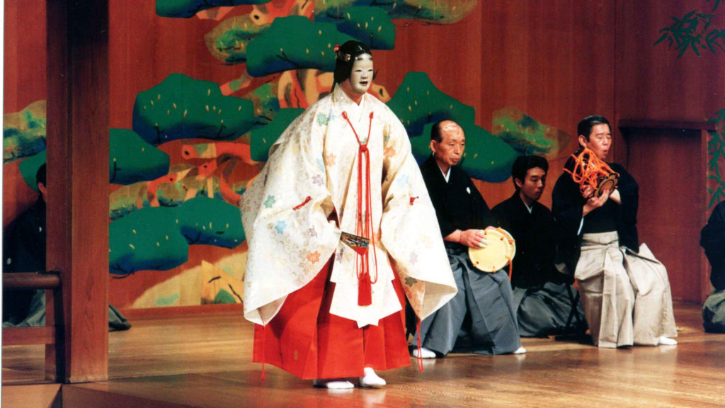 A photo from a performance of a Noh Theatre piece. A masked actor stands centre stage while several musicians sit behind him upstage.