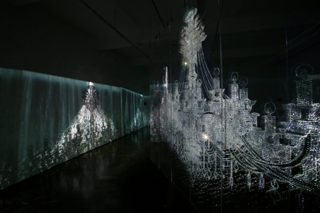 An image of a chandelier work by Ran Hwang. The piece is comprised of an image of a chandelier projected onto a screen, next to an image of a chandelier made from beads and crystals mounted on plexiglass.
