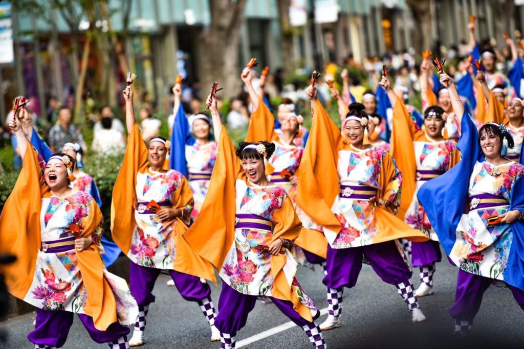 An image of Yosakoi dancers mid-performance on a city street. They are wearing purple and orange costumes and have their arms in the air.