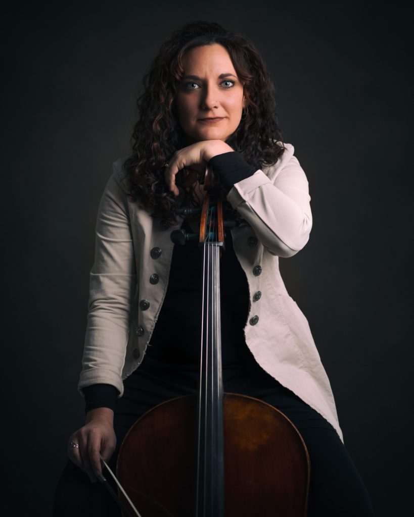 A photo of Aimee Norris posing with her cello. She is seated, looking straight into the camera, resting her hand on the top of her cello, and her head on top of her hand.