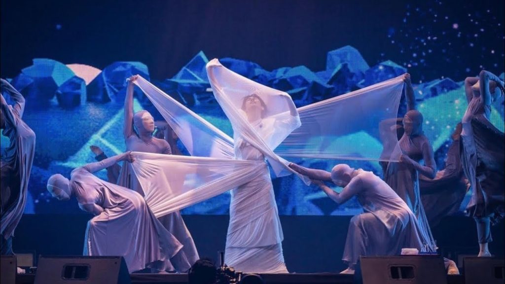 A photo of Build Jakapan onstage, mid-performance with his arms outreached above him. He is wearing all white and is surrounded by dancers in grey who have wrapped him in a white sheer fabric.
