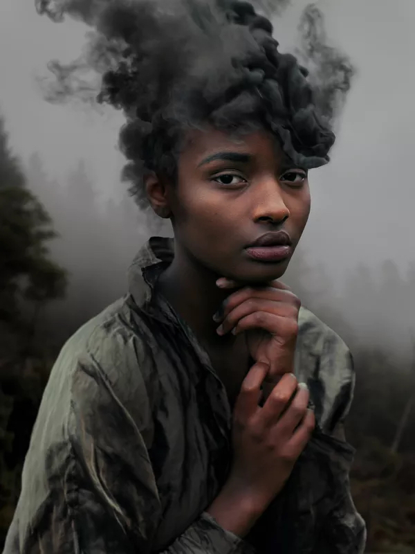 A photography by David Uzochukwu. It shows a woman looking directly into the camera, her hands are held just below her face. Her hair is black smoke above her.
