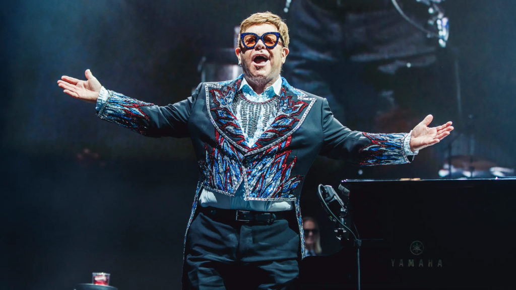 A photo of Elton John onstage during his Farewell Yellow Brick Road Tour. He is facing the audience, holding out his arms to the side, wearing a bedazzled suit.