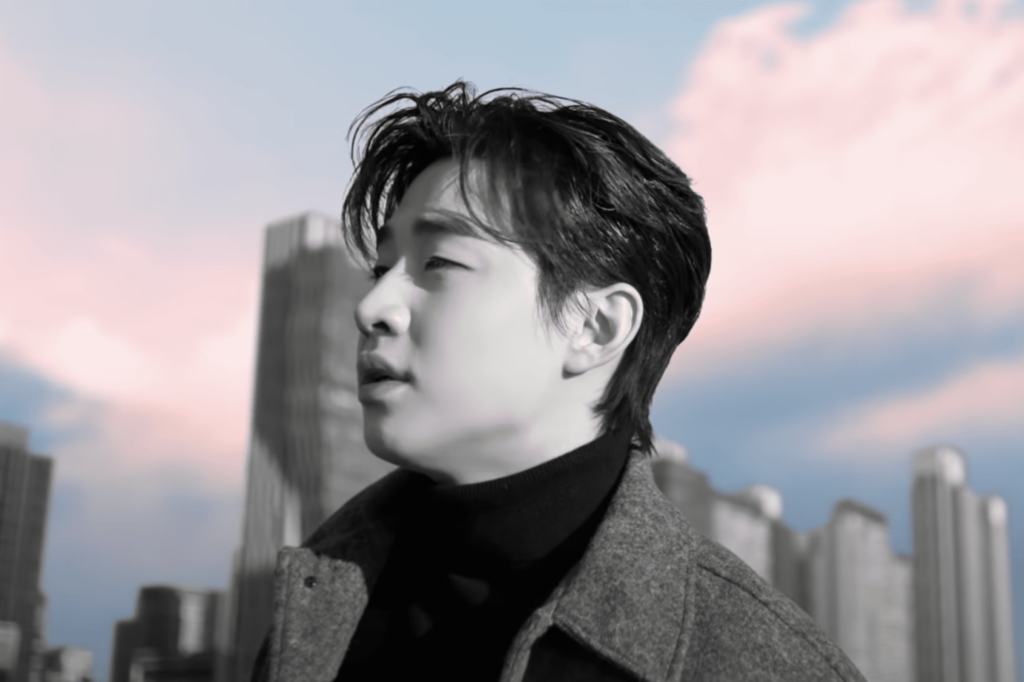 A close up photo of Henry Lau. He is outside, standing in front of buildings, looking off to the left, with a pastel coloured sky behind him.