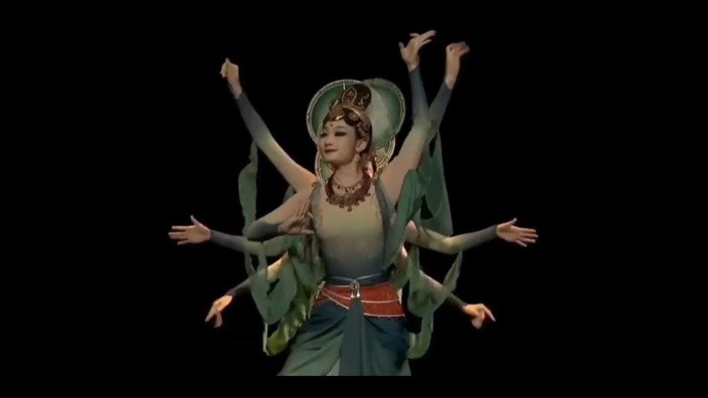 A photo still from a dance piece to Jericho by Iniko. Four dancers are onstage, all standing in a line so that only the front dancer is visible, but with the other dancers' arms sticking out to the sides.