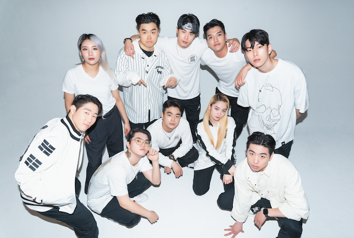 A promotional photo of the Just Jerk Crew. Ten members, all wearing white tops and black pants, are posed in a group looking up at the camera.
