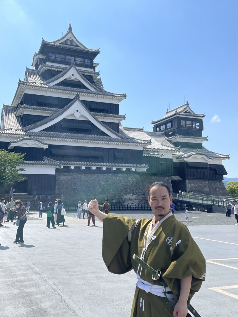 A photo of a man dressed in samurai robes. He is standing in a plaza, holding up one arm and looking right in the camera. The Kumamoto Castle is in the background.