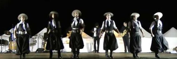 A photo of Malambo dancers. They are standing in a line onstage, wearing traditional outfits and facing the audience.
