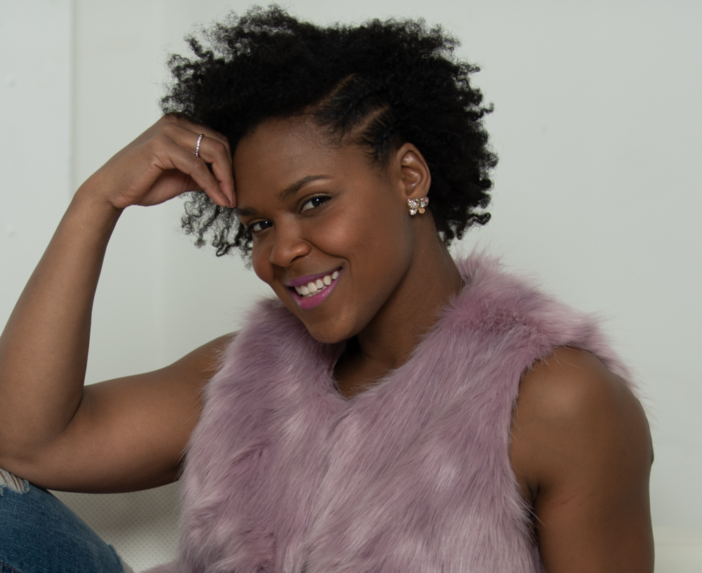 A promotional photo of Shanelle. She is seated with one hand against her head, smiling directly into the camera.