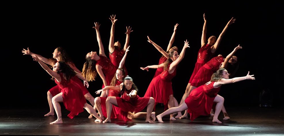 A photo still from the performance of Survivor, choreographed by JudithGilT. 11 dancers in red dresses are mid-performance, their arms outstretched above them in a pose.