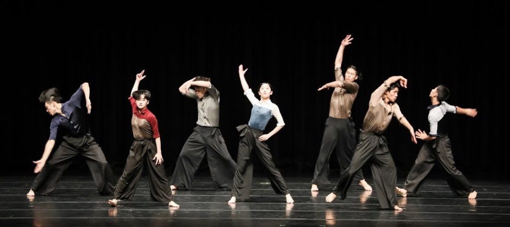 A production photo of Moving Us/Dying Message by Hojeong Choi. Seven performers are standing in a line on a black stage, their hands raised at various levels as they all strike a pose.