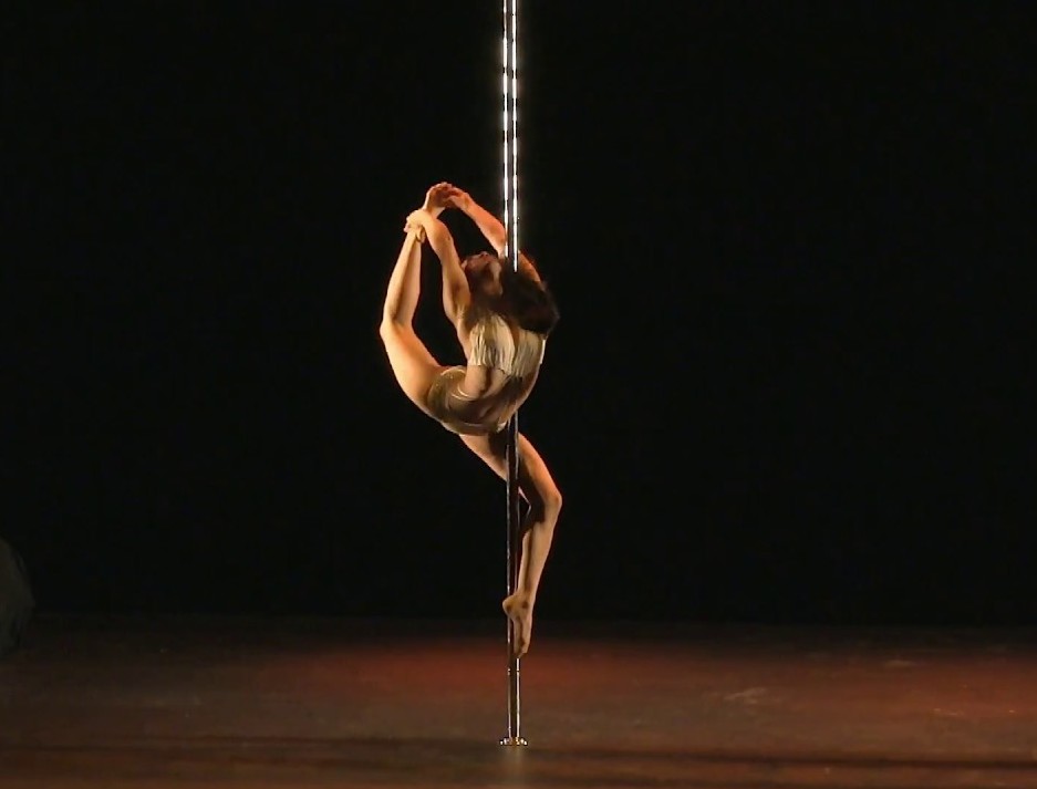 A photo of Renee Wu, mid-performance on a pole. One leg is hooked around the pole and the other is extended up behind her, being held above her head by her arms.