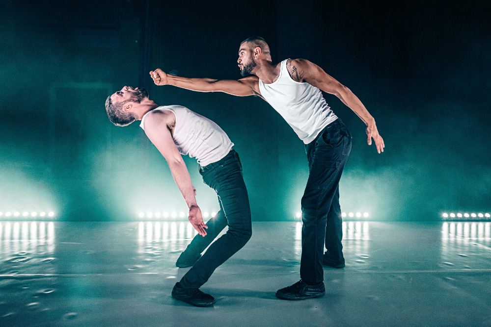 A photo still from Stuntman. Two performers are onstage wearing black pants and white t-shirts. On is punching the other, who is bent backwards to avoid it.