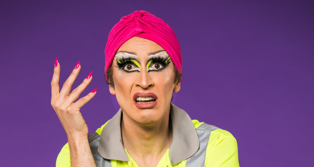 The show image for Wasteman. It is a photo of Joe Leather, wearing a high vis jacket and face made up in drag makeup, looking straight into the camera with one hand raised.