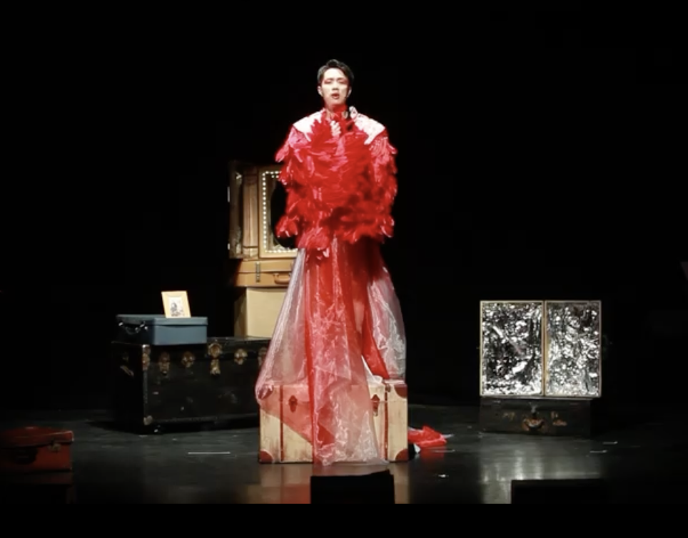 A photo still from Blossoming (You Undo Me). The character of Tao is standing on a box onstage, wearing a red drag outfit.