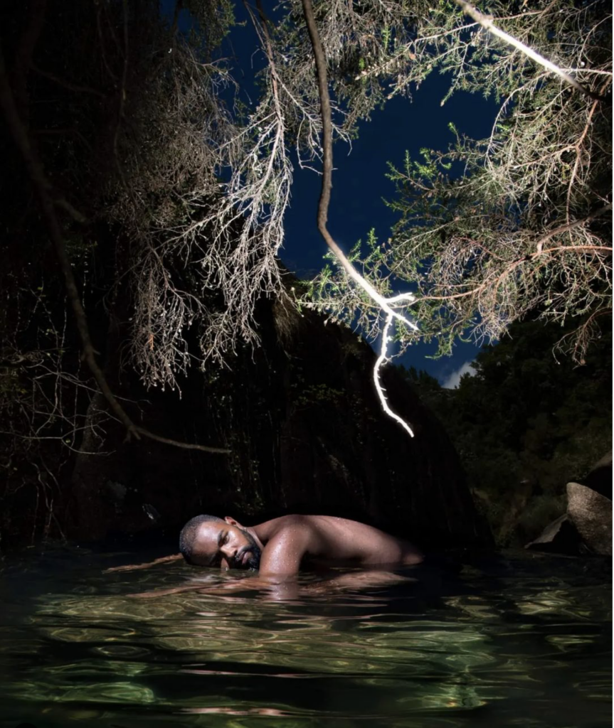 A photograph by Bruno Oliveira. It shows a nude Black man outdoors, bent over in water, looking straight into the camera, with the trees above him illuminated in light.