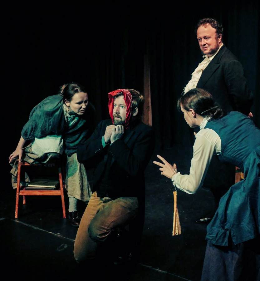 A photo still from a performance of The Grandmothers Grimm. It shows four actors onstage in 19th century costumes. One actor is seated in the centre with a red shawl wrapped around his head, while the other stand around him.