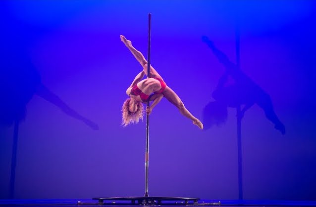 A photo of Laurajane mid-performance on a pole. Her body is horizontal and her legs are outstretched in the splits.