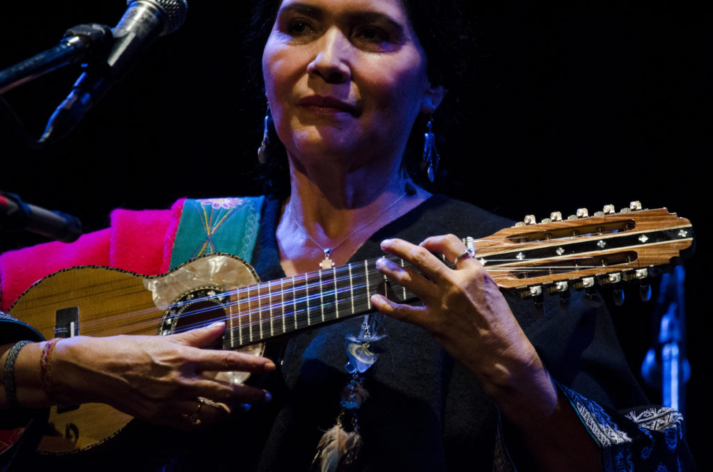 A close up photo of Luzmila Carpio onstage performing. She is standing in front of a microphone and holding a guitar.