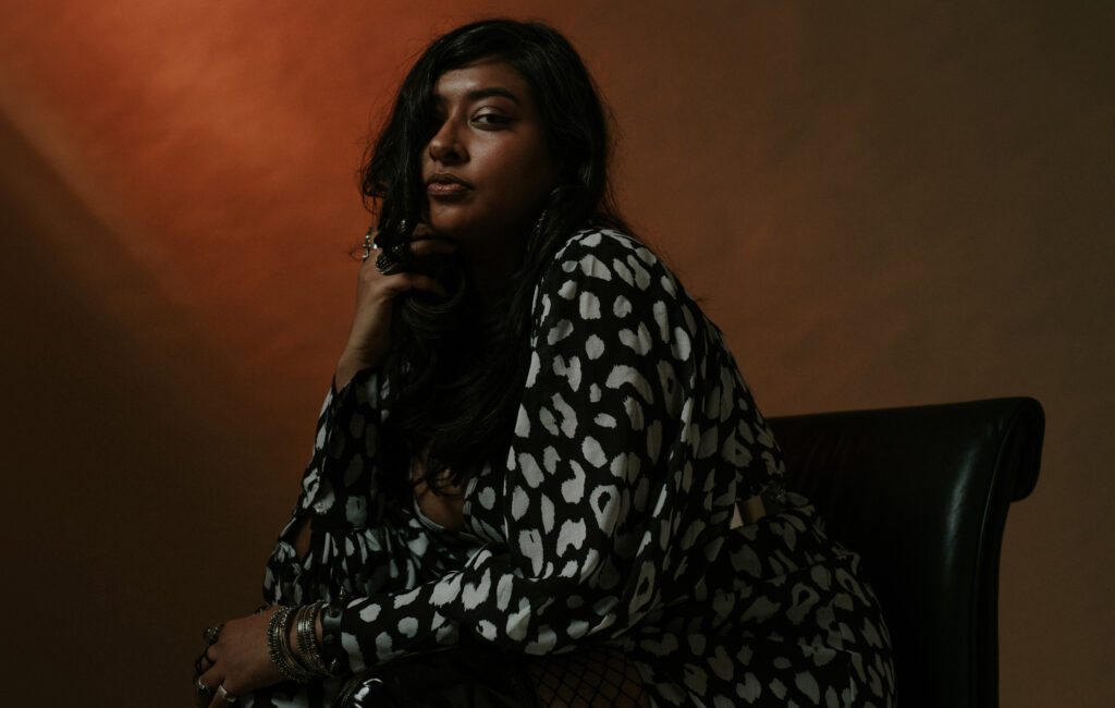 A promotional photo of Meghna Jayanth. She is sitting in profile, her head turned towards the camera, with a dark orange background behind her.