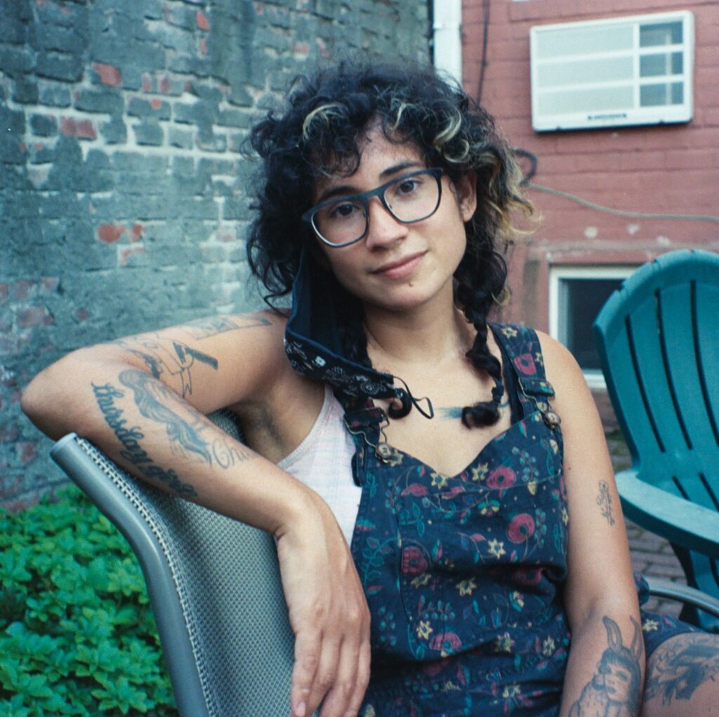 A promotional photo of Melissa Lozada-Oliva. She is sitting sideways on a chair, one arm up on the chair back, looking straight into the camera.