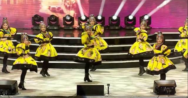 A photo of Romanian children dressed in yellow dresses, mid-dance performance on a stage with a pink background.
