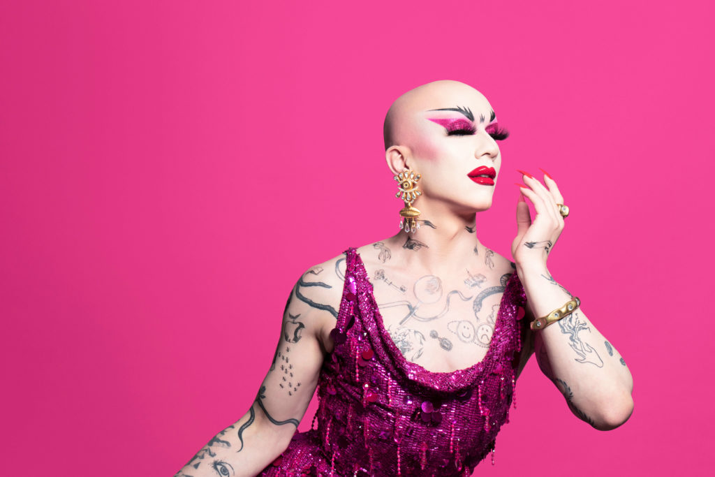 A photo of Sasha Velour. She is dressed in a pink dress, posing in front of a bright pink wall.