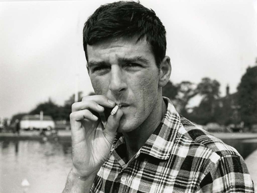 A black and white candid photo of Thom Gunn. His is smoking a cigarette, looking straight into the camera.