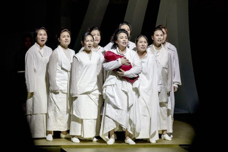A photo still from The Trojan Women. The eight women chorus are standing in a group around Hecuba. They all wear white, and Hecuba in the centre is holding a red cloth wrapped baby bundle.