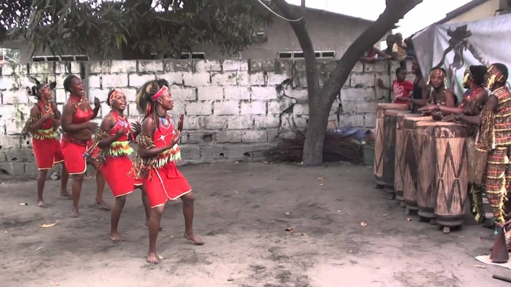 A photo still from a performance by Ballet Arumbaya Ndendeli. Several men are playing instruments on the right side of the stage, while four women dance in a line on the left side of the stage.