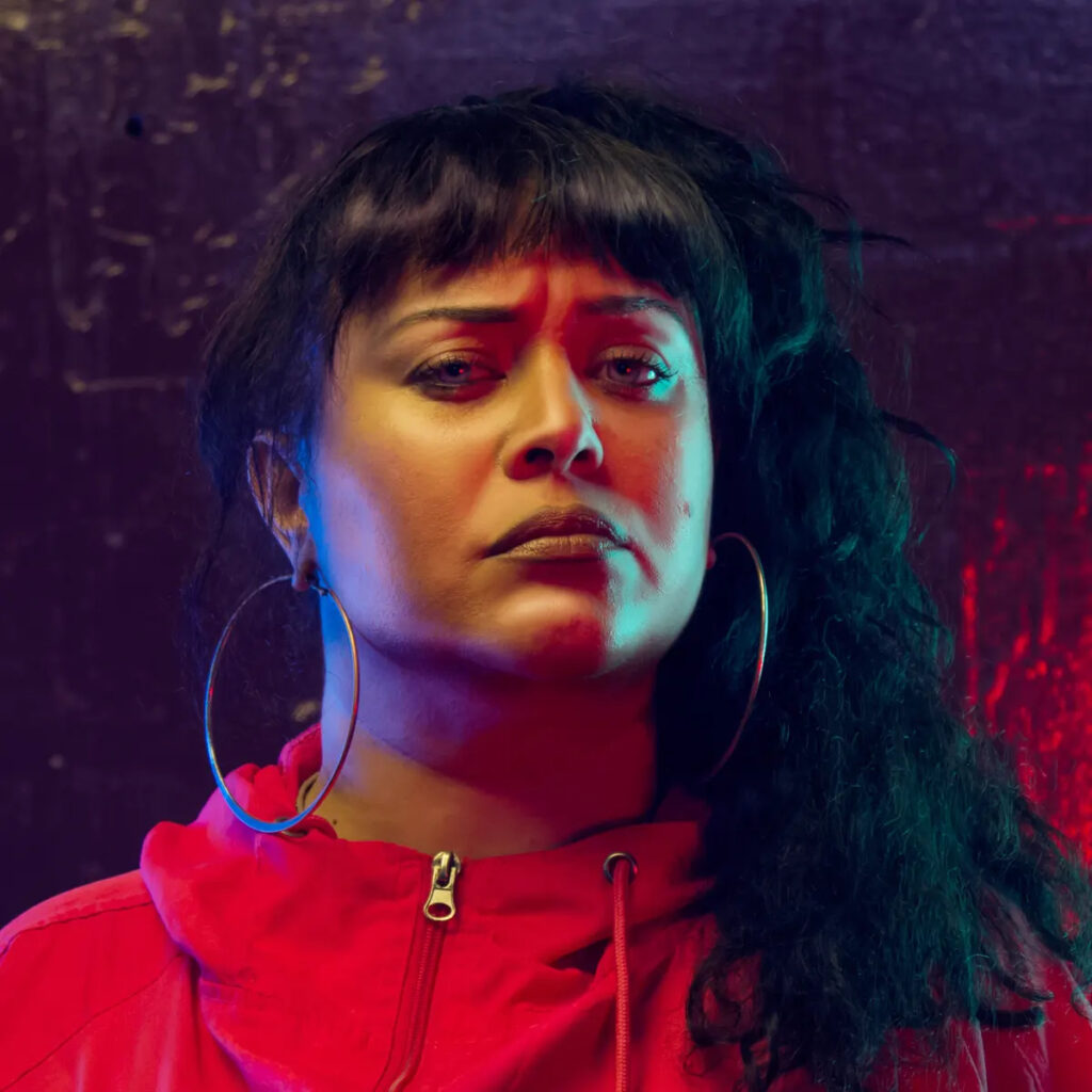 A promotional headshot of Coco Solid. She is looking straight into the camera, wearing a red top against a dark purple background, and a neutral expression on her face.