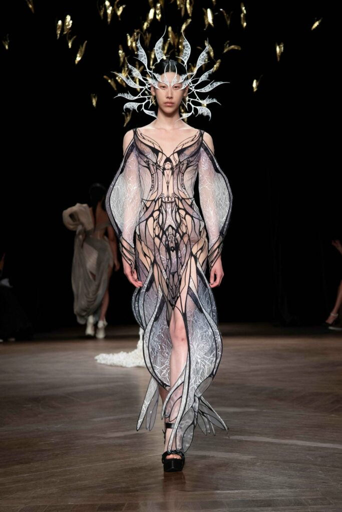 A photo of an outfit designed by Iris van Herpen. The model is wearing a billowing dress with flared sleeve and a crown on their head.