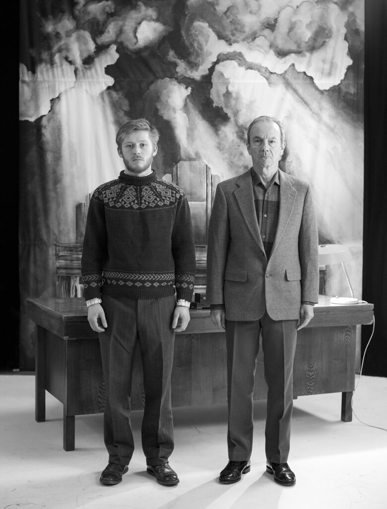 A black and white photo from the art installation NSFW: A Chairman's Tale by Jannus Samma. It shows two men, one young one older, standing stiffly side by side in front of an office desk.