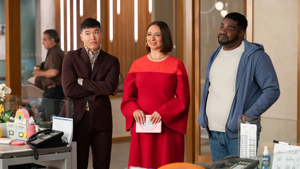 A photo still from Loot. Maya Rudolph, Joel Kim Booster, and Ron Funches are standing in a line in character, looking just off camera at an unseen character.